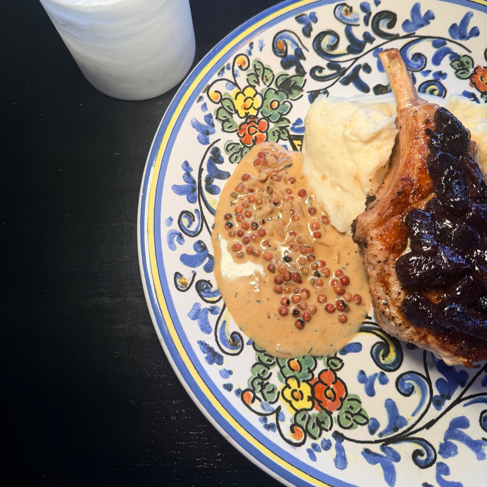 Roasted duck leg with blackberry sauce, peppercorn cream, and mashed potatoes on a colorful patterned plate beside a glass of water.