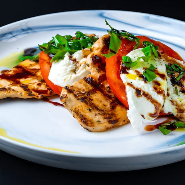 Grilled chicken with sliced tomatoes, fresh mozzarella, basil, and balsamic glaze on a white plate.