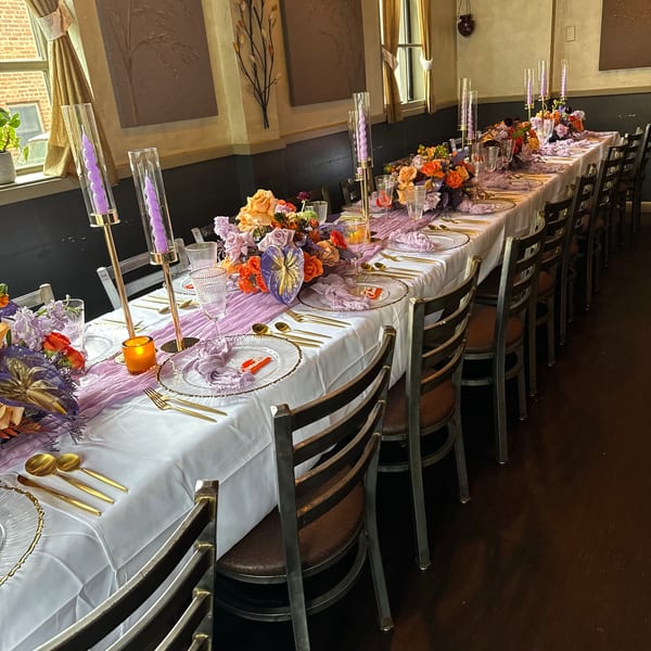 Long dining table set with purple and orange floral arrangements, candles, and gold flatware at The 808 Bistro.