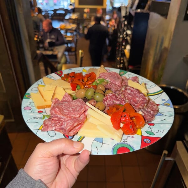 A hand holds a colorful plate of Italian charcuterie with meats, cheeses, olives, and roasted peppers.