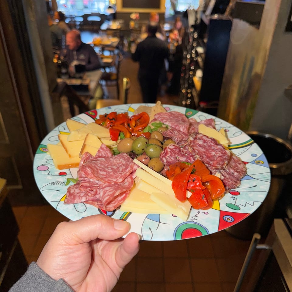 A hand holds a colorful plate of Italian charcuterie with meats, cheeses, olives, and roasted peppers.