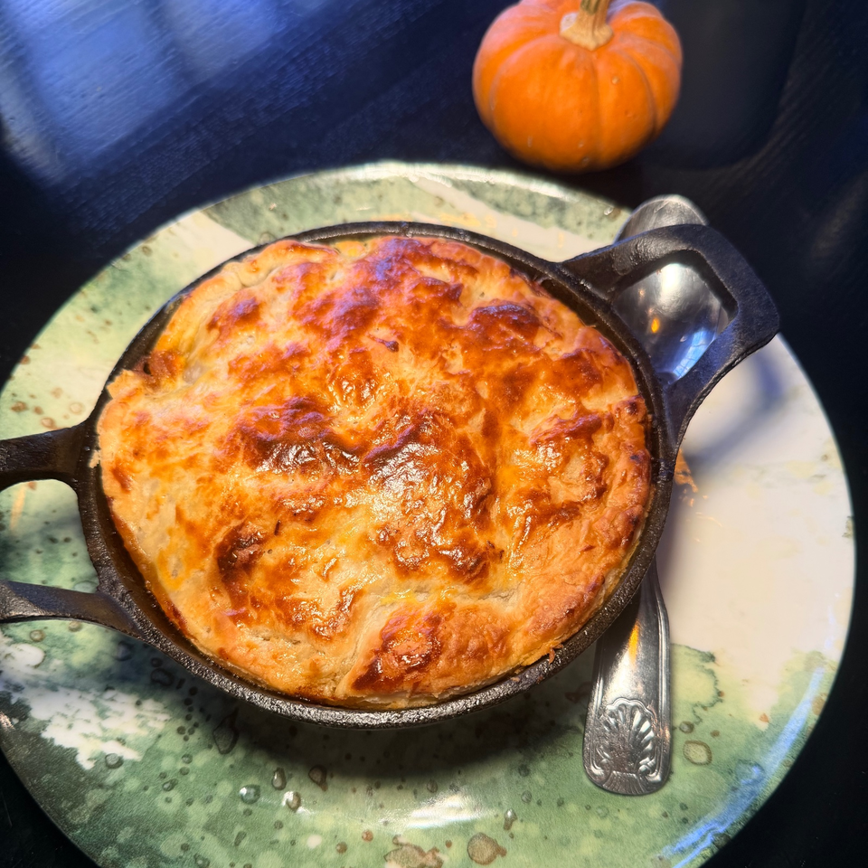 Golden-brown cheese-topped savory pie in a cast-iron skillet on a decorative plate, with a small pumpkin and spoon nearby.